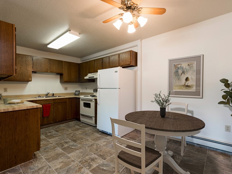 Bismarck, ND Rosser Apartments. A dining room with a table and two chairs. The overhead fan and light brighten the room. A kitchen with a white refrigerator freezer next to a white stove top oven are in the background